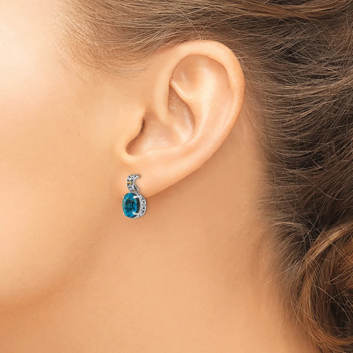A person wearing a teardrop-shaped earring with a blue stone surrounded by smaller diamonds or crystals in a white metal setting.