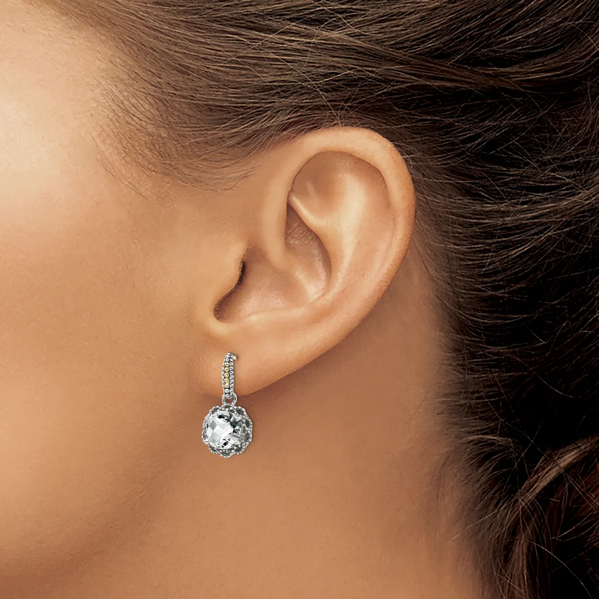 A woman wearing a silver drop earring with a round clear gemstone surrounded by smaller diamonds, shown from the side view of her ear.