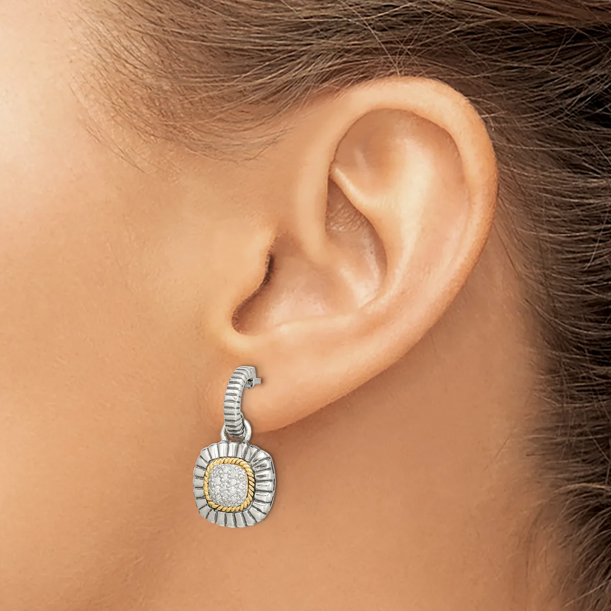 A woman wearing a two-tone drop earring with a cushion-shaped white diamond or gemstone surrounded by a fluted metal halo and attached to a textured post.