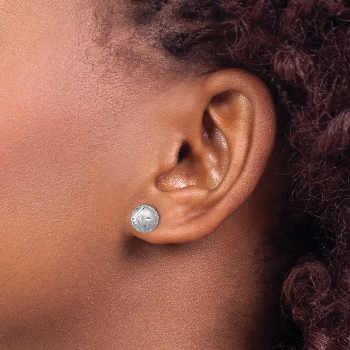 A person wearing a small round silver stud earring with a textured surface in their earlobe, shown in profile against their neck and shoulder.