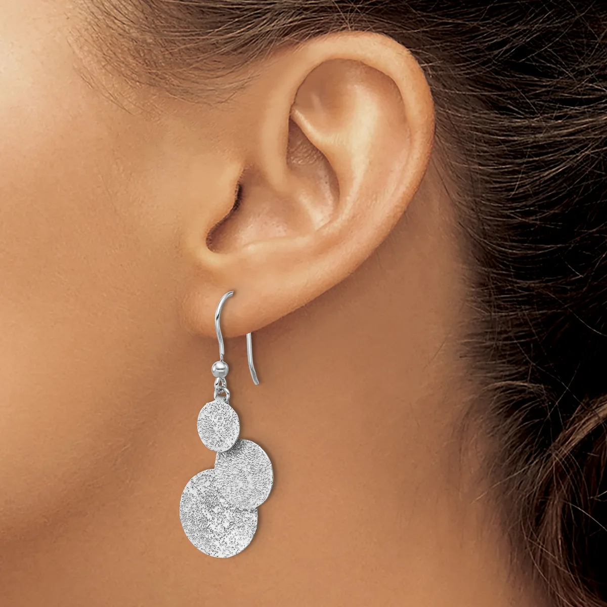 A woman wearing a pair of silver drop earrings with two connected circular pendants that have a textured finish, suspended from curved ear wires.