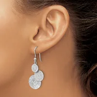 A woman wearing a pair of silver drop earrings with two connected circular pendants that have a textured finish, suspended from curved ear wires.