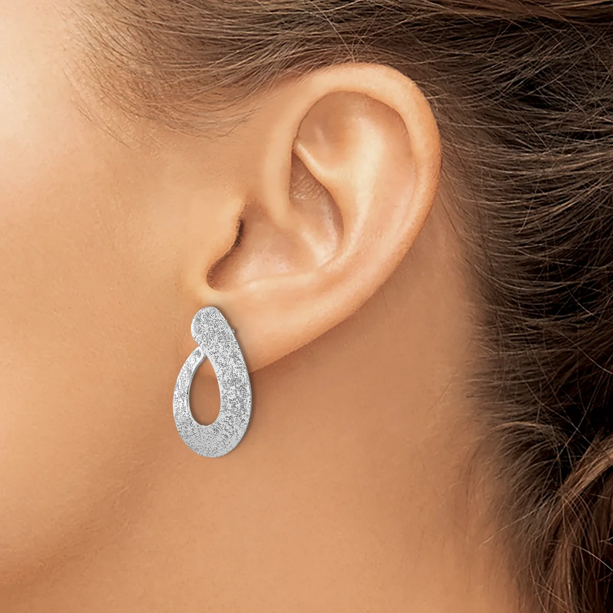 A woman wearing a textured silver teardrop-shaped hoop earring with an open center, shown from the side view of her ear.