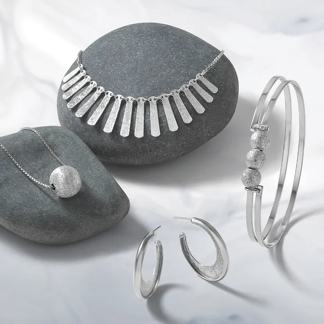 Four pieces of silver jewelry displayed on gray stone: a necklace with rectangular pendants, a pendant necklace with a textured sphere, a bangle bracelet with three textured spheres, and a pair of hoop earrings with textured finish.