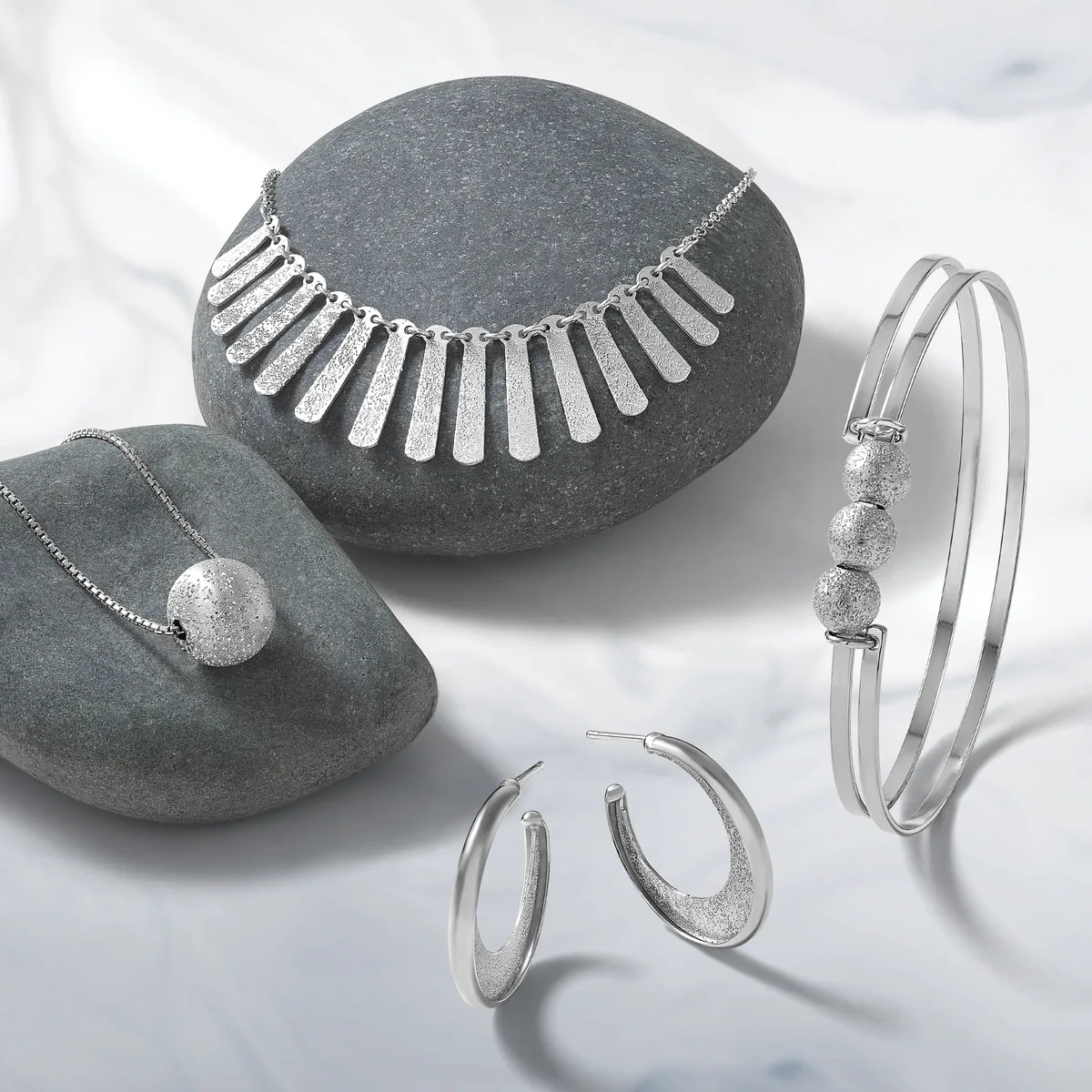 Four pieces of silver jewelry displayed on gray stone: a necklace with rectangular pendants, a pendant necklace with a textured sphere, a bangle bracelet with three textured spheres, and a pair of hoop earrings with textured finish.