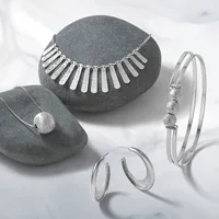 Four pieces of silver jewelry displayed on gray stone: a necklace with rectangular pendants, a pendant necklace with a textured sphere, a bangle bracelet with three textured spheres, and a pair of hoop earrings with textured finish.
