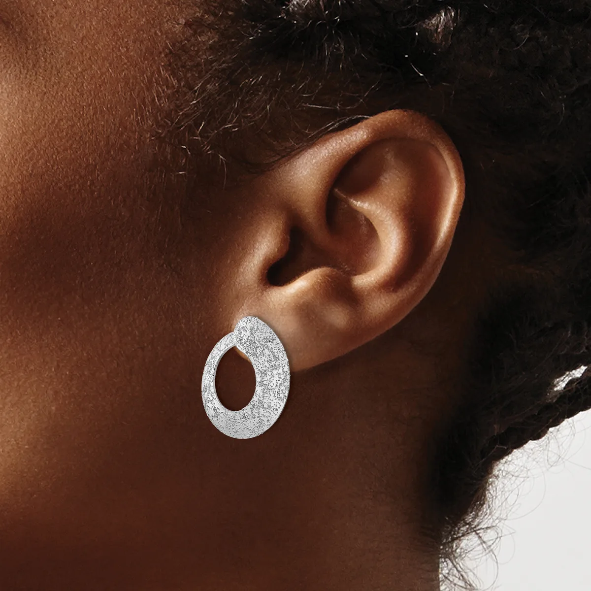 A person wearing a textured silver oval hoop earring with an open center, shown from the side profile against their ear.
