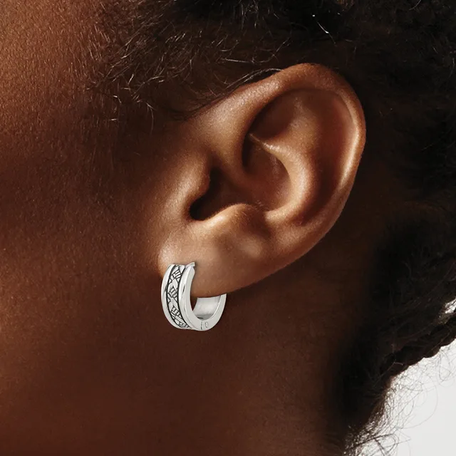 A silver hoop earring with an engraved decorative band worn on the earlobe of a person with brown skin.