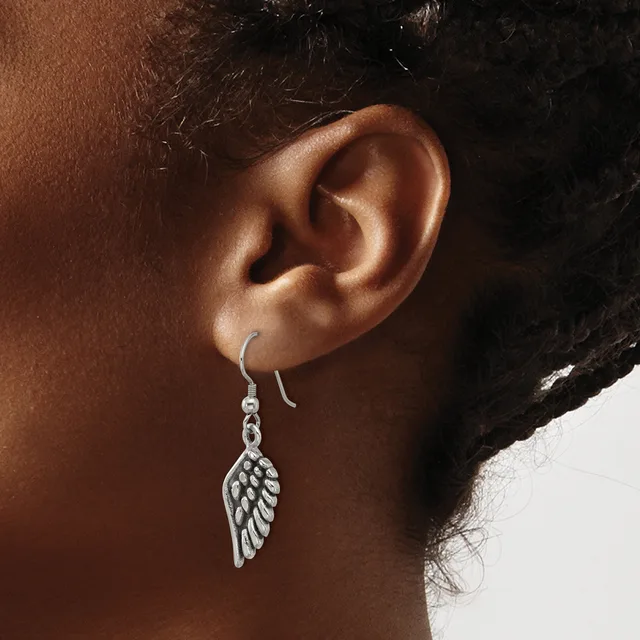 A silver wing-shaped dangle earring hangs from a curved hook on a person's earlobe.