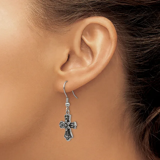 A person wearing a silver-toned cross earring with ornate detailing, shown hanging from the ear lobe against their shoulder.