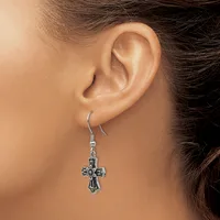 A person wearing a silver-toned cross earring with ornate detailing, shown hanging from the ear lobe against their shoulder.