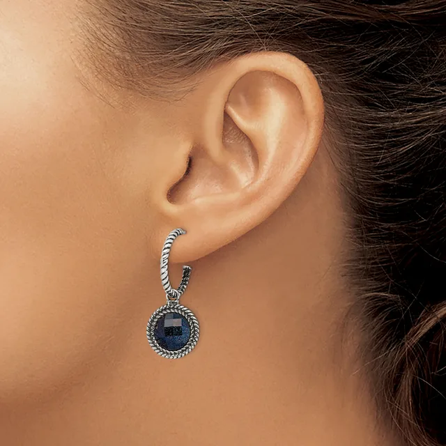 A woman wearing a silver-toned drop earring with a twisted rope-textured top loop and a round dark blue stone pendant with a beaded border, shown from the side profile.