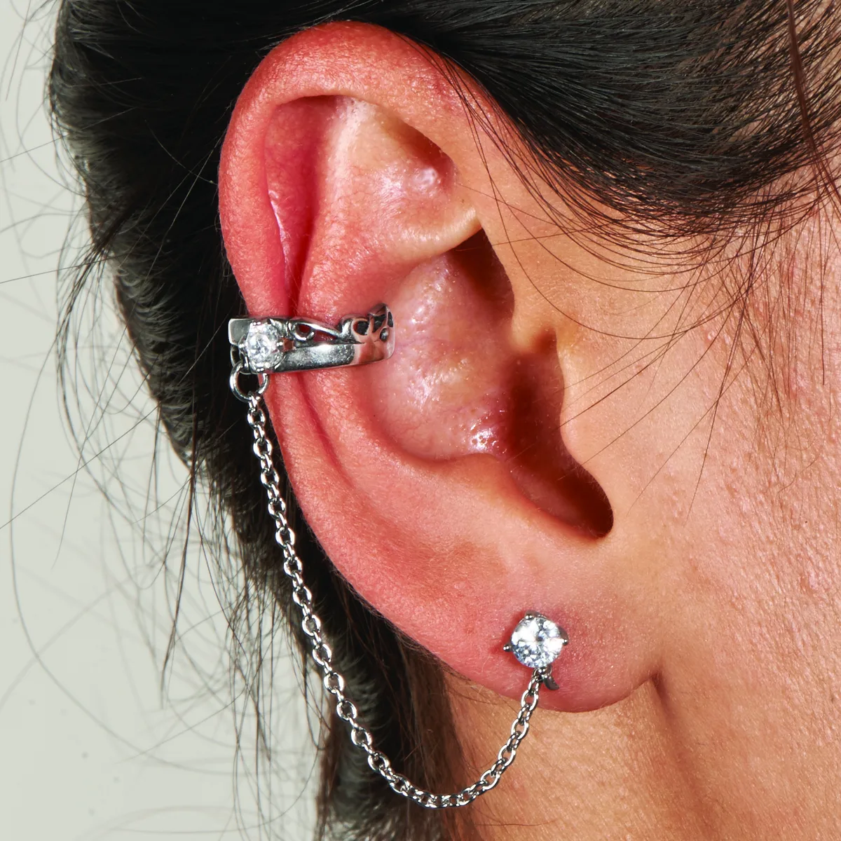 A person wearing a silver-toned ear cuff with a decorative knot design on the upper ear, connected by a chain to a stud earring with a round clear stone in the lower lobe.
