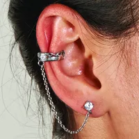 A person wearing a silver-toned ear cuff with a decorative knot design on the upper ear, connected by a chain to a stud earring with a round clear stone in the lower lobe.