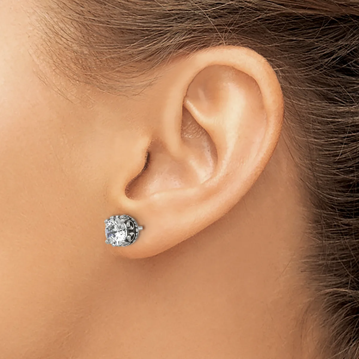 A close-up side view of a person's ear wearing a silver stud earring with a round clear gemstone in an ornate setting.