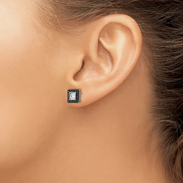 A square stud earring with a clear center stone surrounded by dark metalwork sits on the earlobe of a person shown in profile.