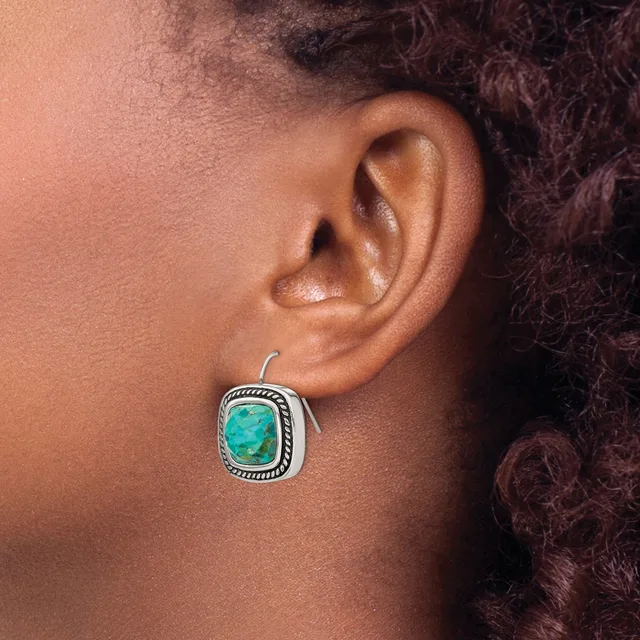 A person wearing a silver square earring with a turquoise stone and decorative beaded border, shown on their ear from the side.