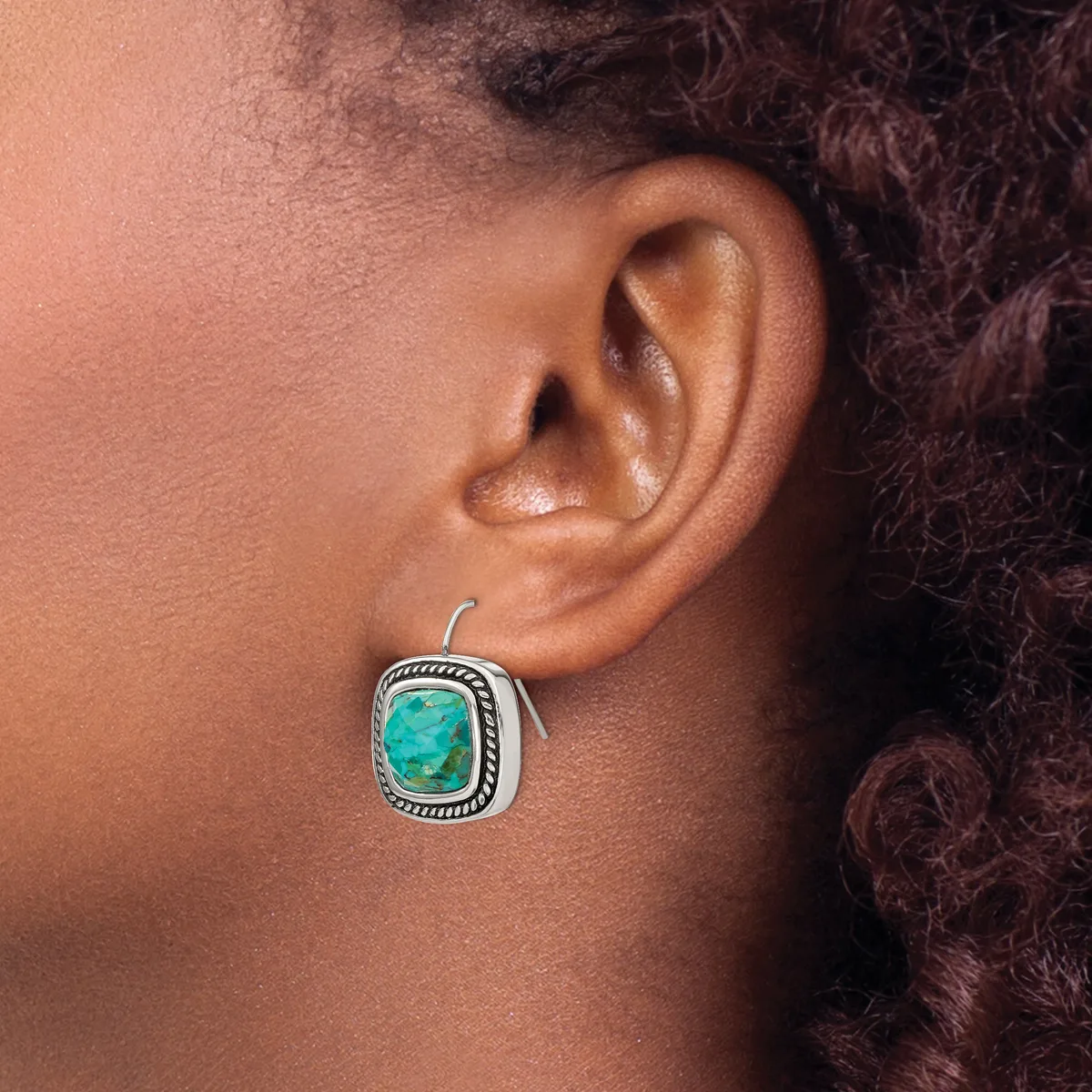A person wearing a silver square earring with a turquoise stone and decorative beaded border, shown on their ear from the side.