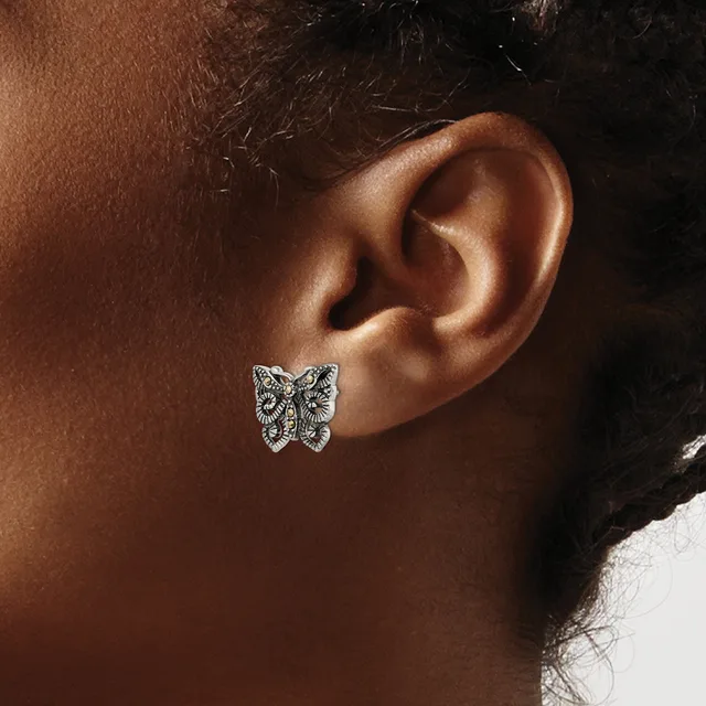 A butterfly-shaped stud earring with intricate filigree detailing worn on the earlobe of a person with dark skin and black hair.