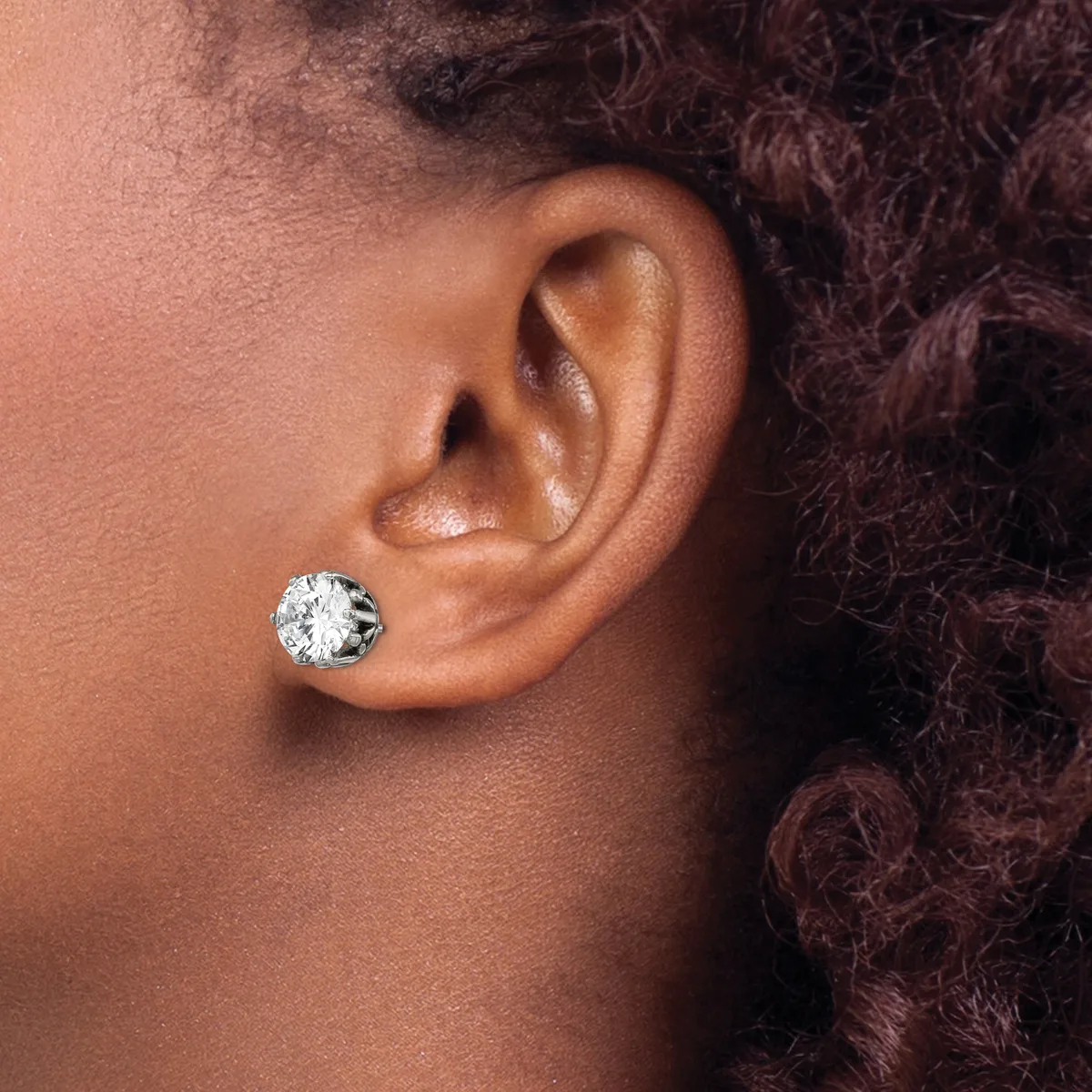 A person wearing a round diamond stud earring in their pierced earlobe, shown in profile against their shoulder and dark curly hair.