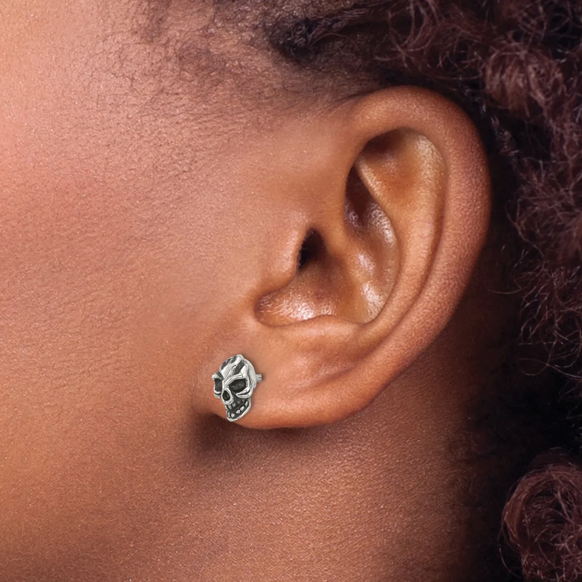 A silver skull-shaped earring stud worn in the earlobe of a person with medium skin tone and dark curly hair.