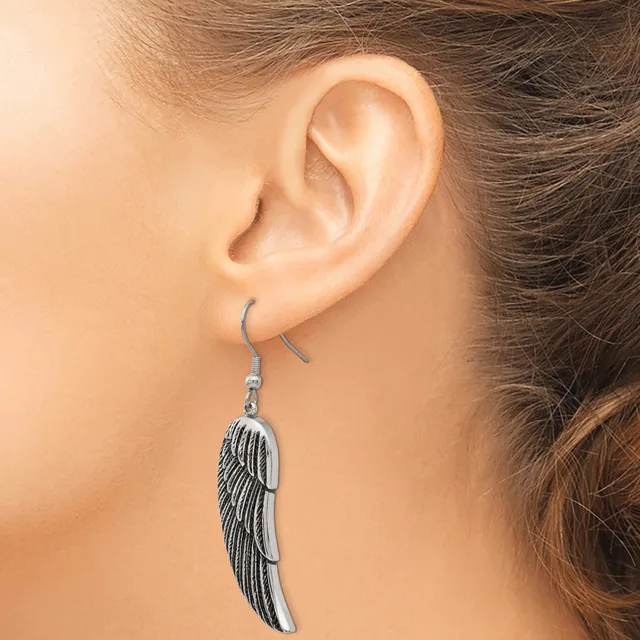 A silver-toned feather-shaped dangle earring hangs from a curved wire hook on a person's earlobe.