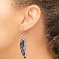 A silver-toned feather-shaped dangle earring hangs from a curved wire hook on a person's earlobe.