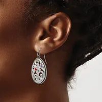 A person wearing a teardrop-shaped silver earring with ornate scrollwork and red gemstone accents, shown hanging from the earlobe against skin and dark hair.