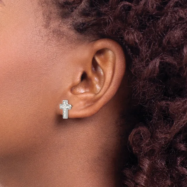 A silver cross earring with an outlined design is worn in the earlobe of a person with brown skin and dark curly hair.