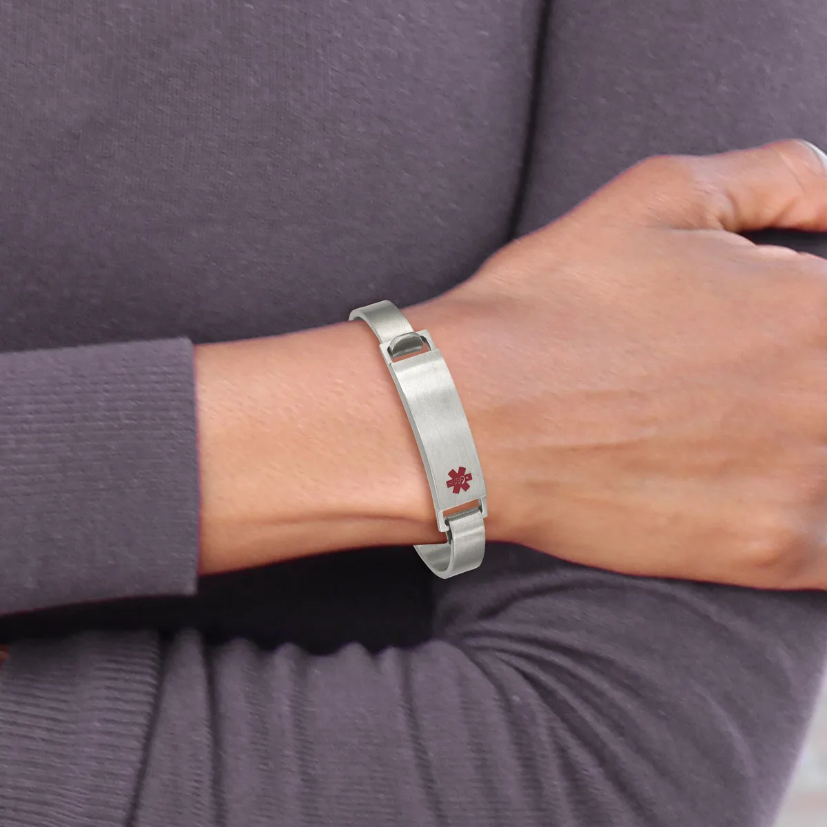 A person wearing a purple long-sleeved shirt displays a silver bangle bracelet with a small red medical alert symbol on their wrist.