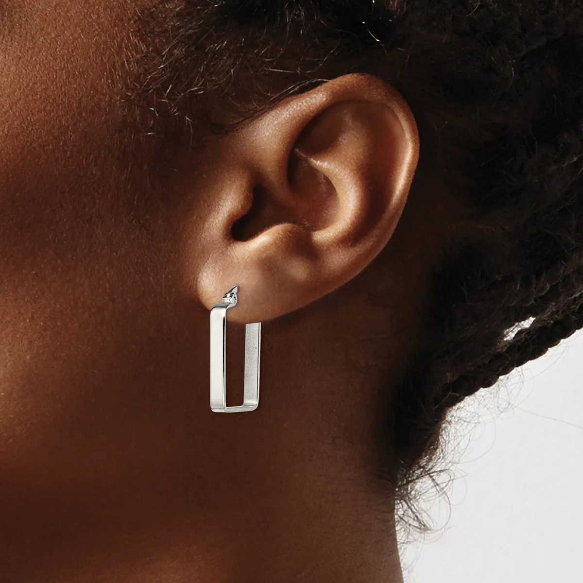 A person wearing a rectangular silver hoop earring with an open center design, shown from the side view against their ear.