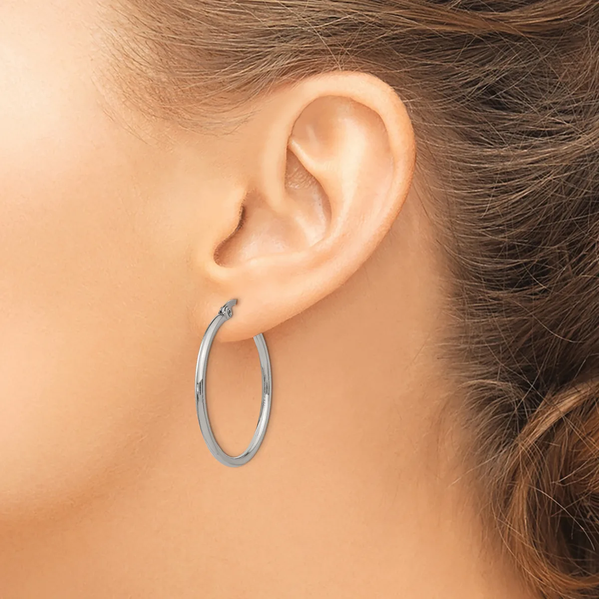 A person wears a pair of polished oval hoop earrings in silver-toned metal, shown from a side profile view of their ear.