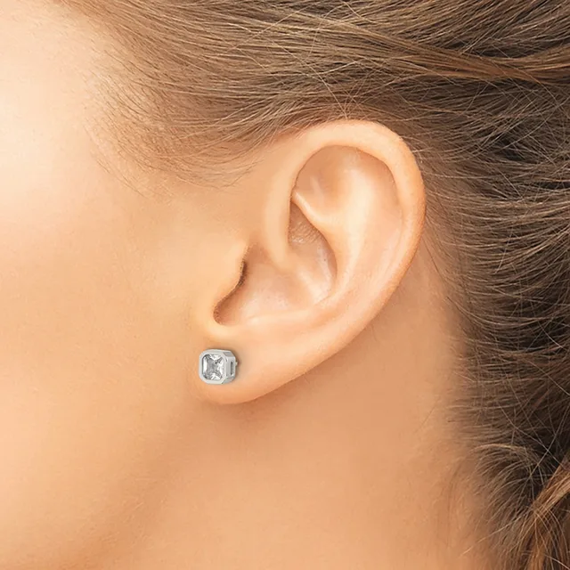 A person wearing a small square stud earring with a clear gemstone set in a metallic bezel in their earlobe.