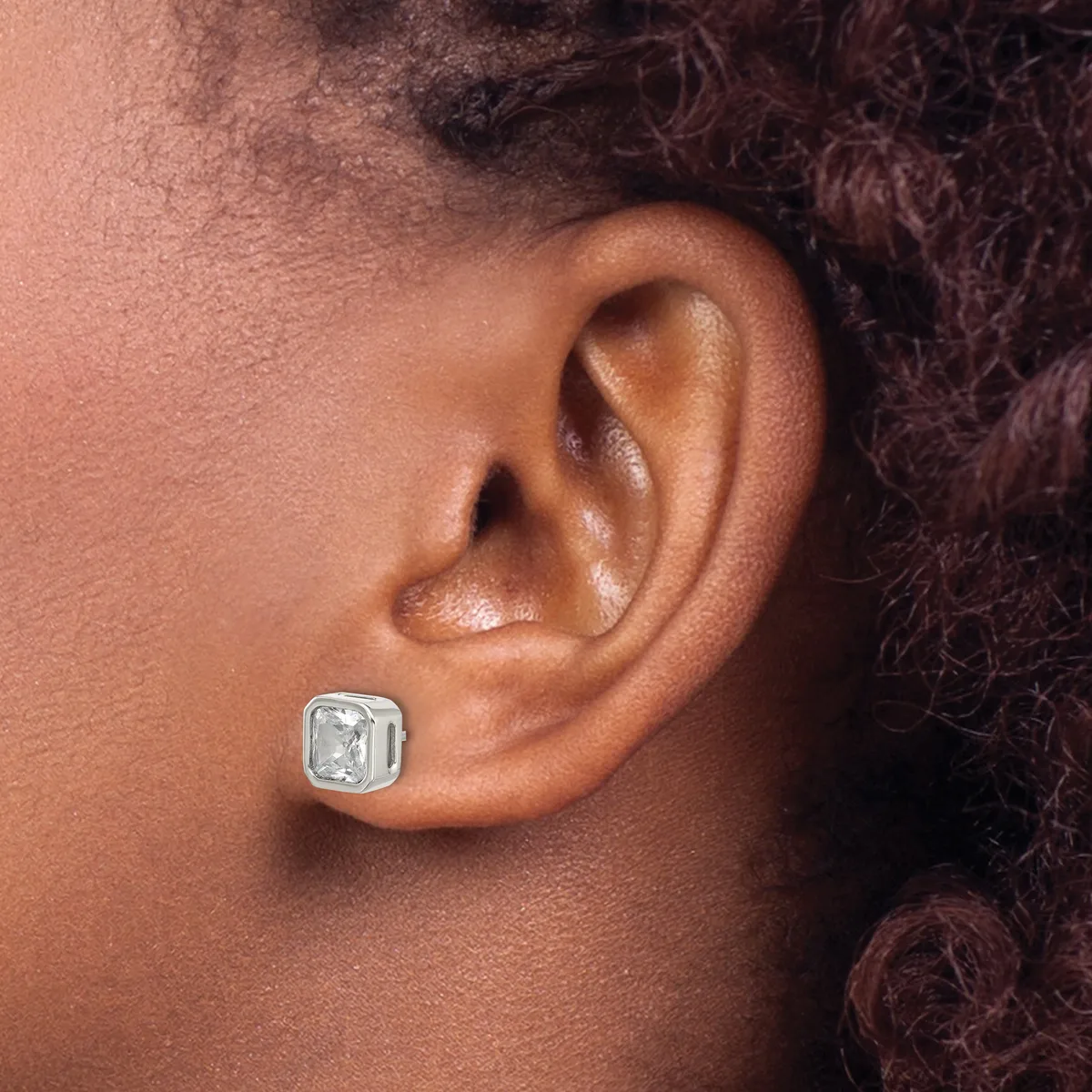 A silver square stud earring with a clear rectangular stone sits on the earlobe of a person with brown skin and dark curly hair.