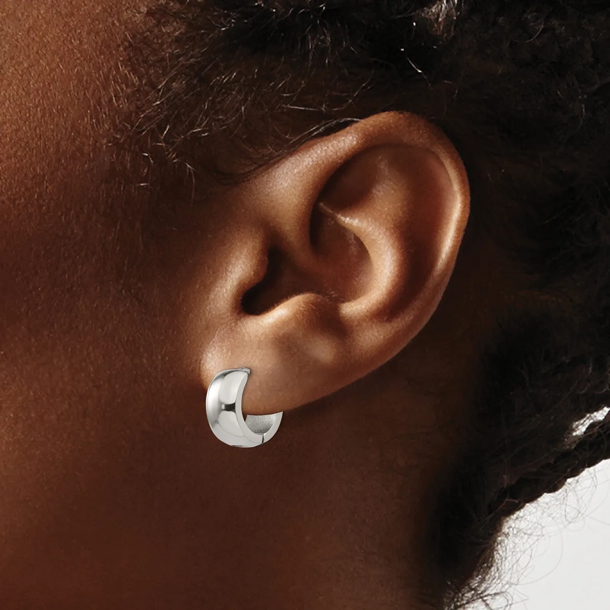 A polished silver chunky curved hoop earring worn on the earlobe of a person with medium brown skin tone and dark hair.