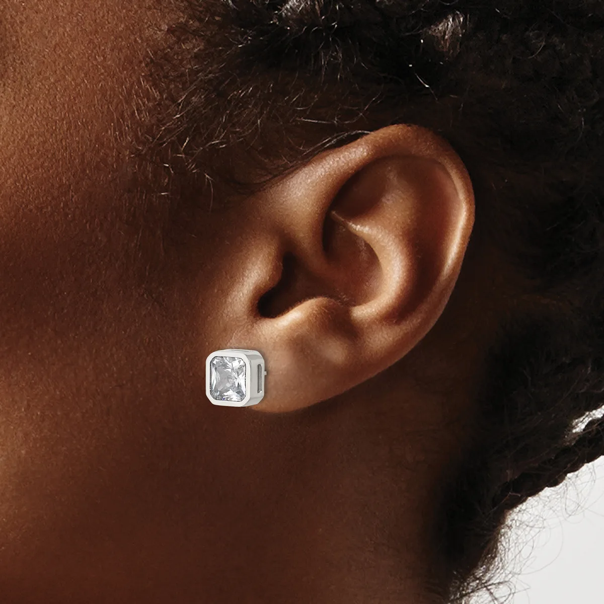 A silver square stud earring with a rectangular white gemstone sits on a person's earlobe.
