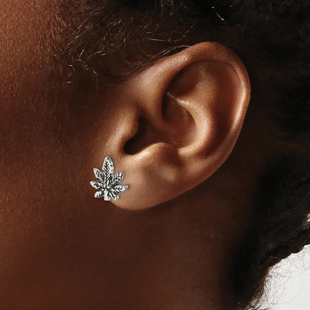 A silver leaf-shaped stud earring with textured detailing worn on the earlobe of a person with brown skin.