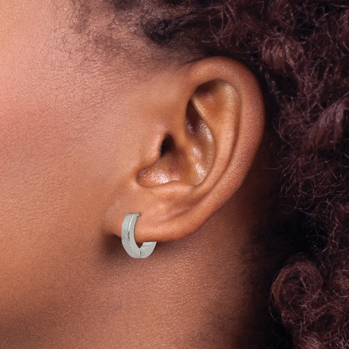 A person wearing a textured silver-toned hoop earring in their earlobe, shown in profile against their skin.