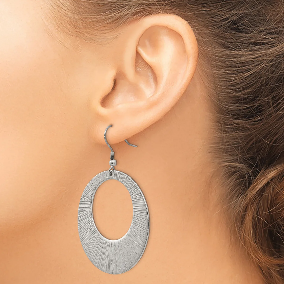 A woman wearing a silver-toned oval dangle earring with a textured brushed finish, shown from the side view of her ear.