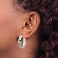 A person wearing a pair of silver hoop earrings with a textured wavy pattern on one side and smooth finish on the other, shown from the side profile.