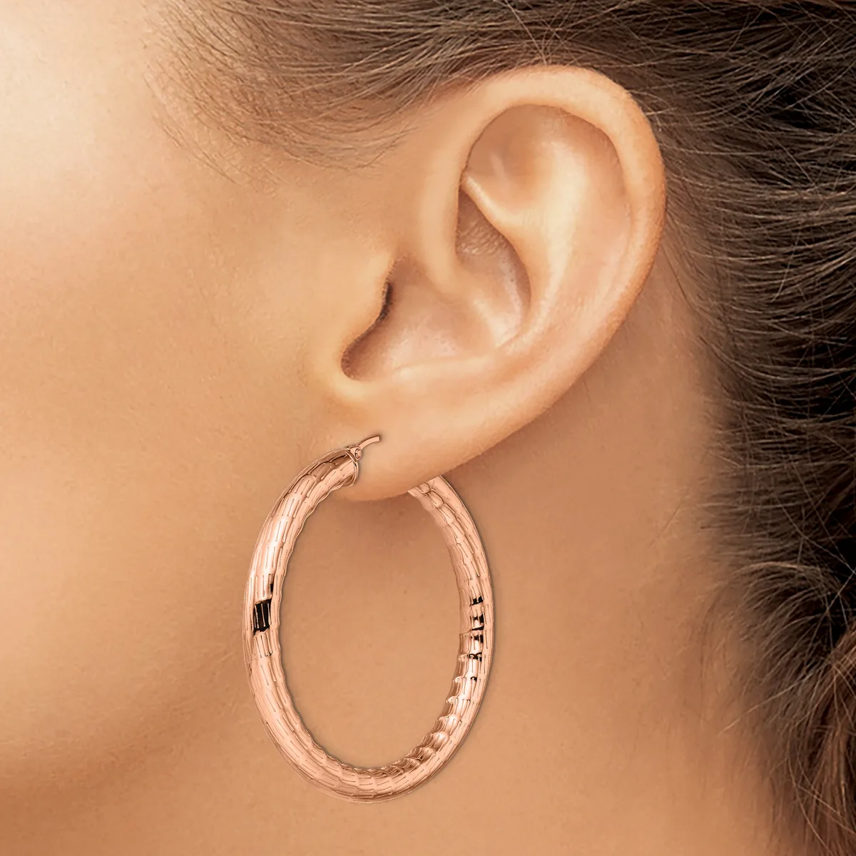 A woman wearing a rose gold textured oval hoop earring, shown from the side profile of her ear and neck.