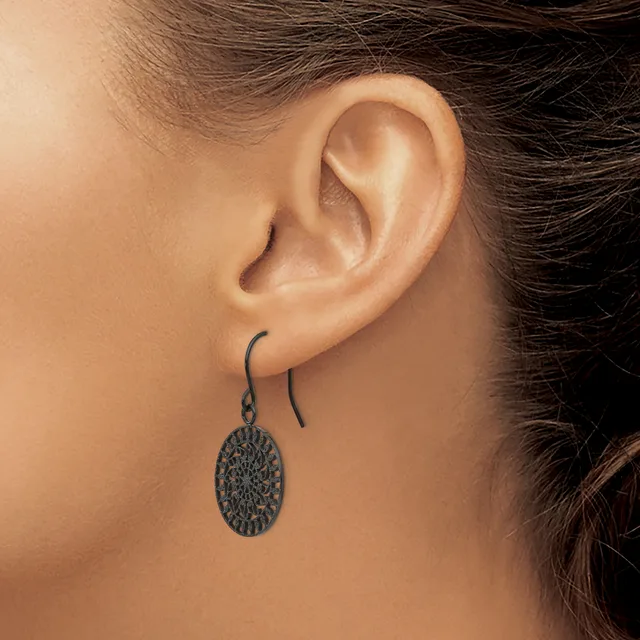 A person wearing a pair of dark metal oval dangle earrings with a textured surface on curved ear wires.
