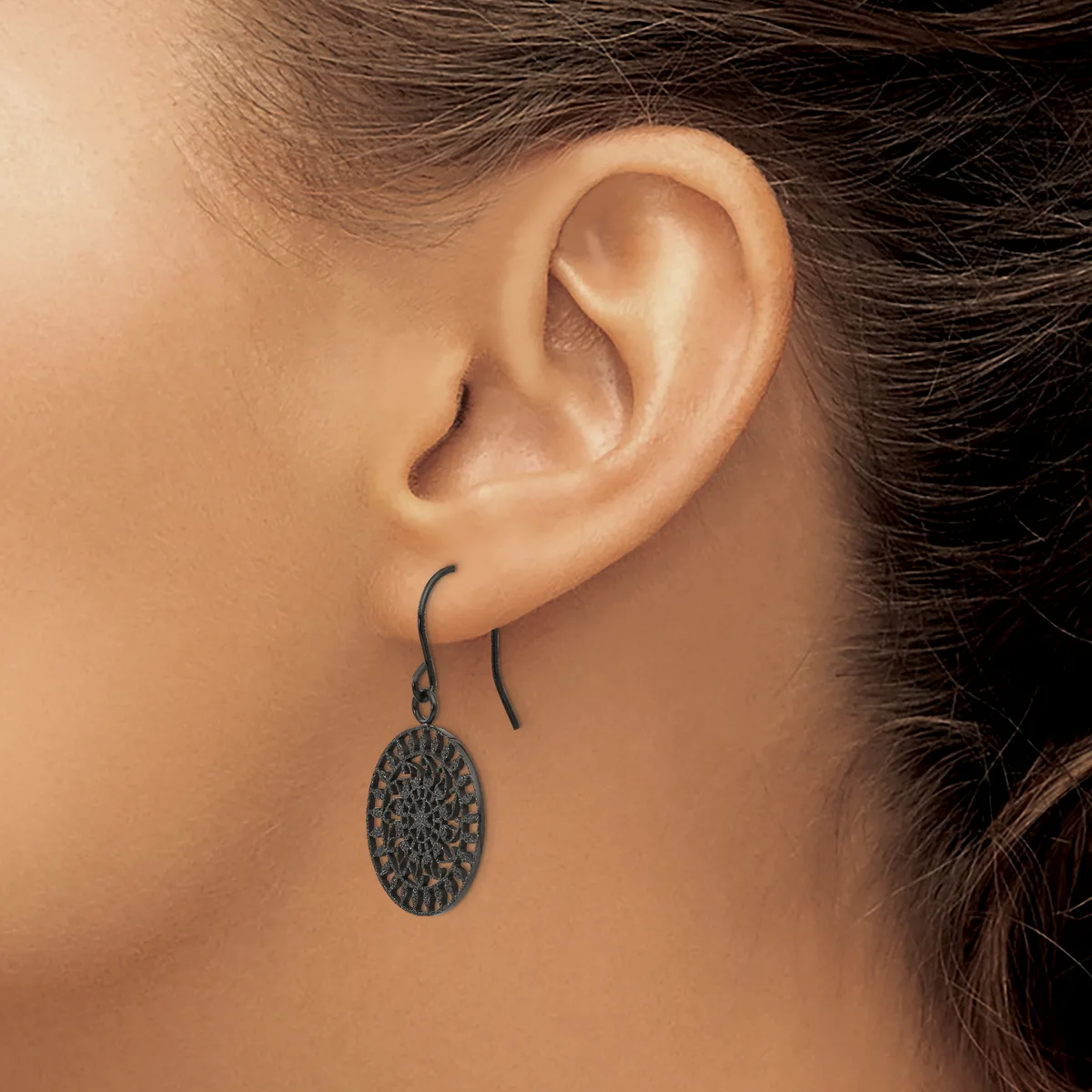 A person wearing a pair of dark metal oval dangle earrings with a textured surface on curved ear wires.