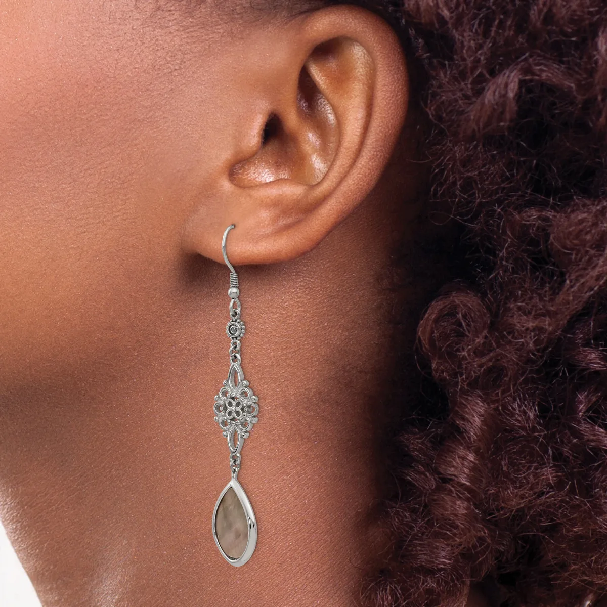 A silver filigree drop earring with an ornate diamond-shaped top and teardrop-shaped stone pendant, worn on a person with dark curly hair.