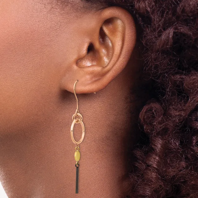 A person wearing a dangling earring with a gold oval link at the top, a small yellow-green stone in the middle, and a dark rectangular charm hanging below.