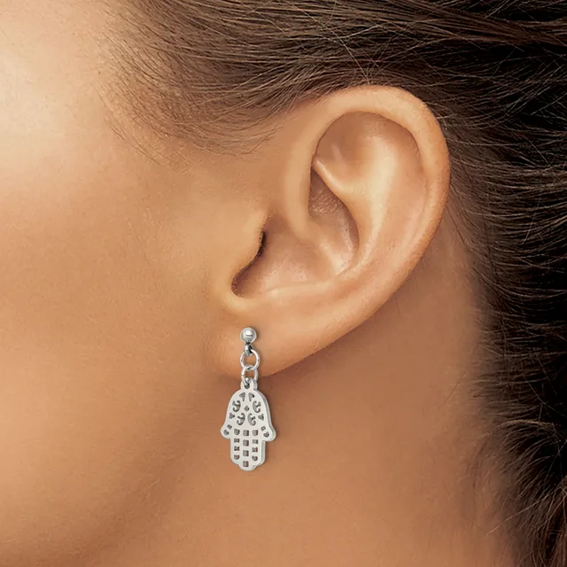 A silver hamsa hand charm earring with decorative cutout details hangs from a stud on a person's earlobe.