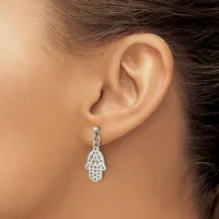 A silver hamsa hand charm earring with decorative cutout details hangs from a stud on a person's earlobe.