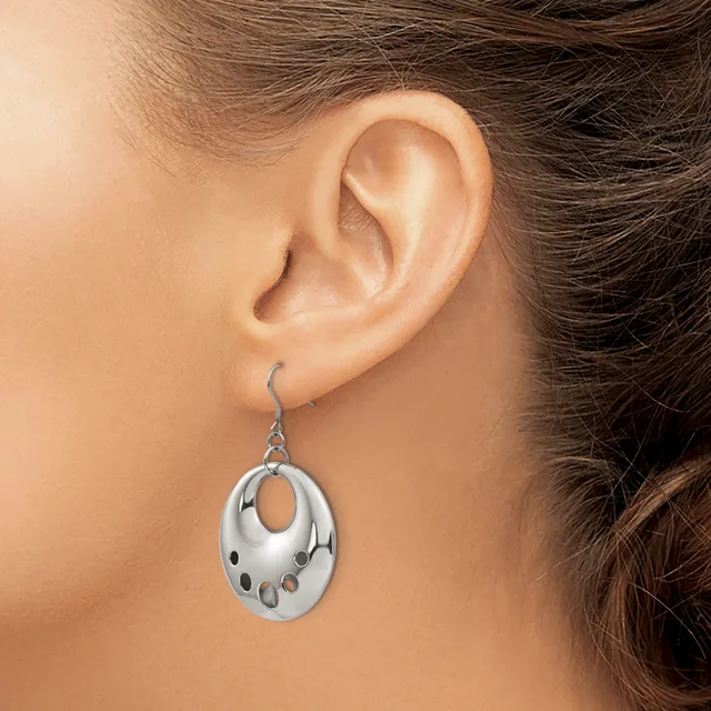 A silver oval pendant earring with cutout details hangs from a wire hook on a person's earlobe.