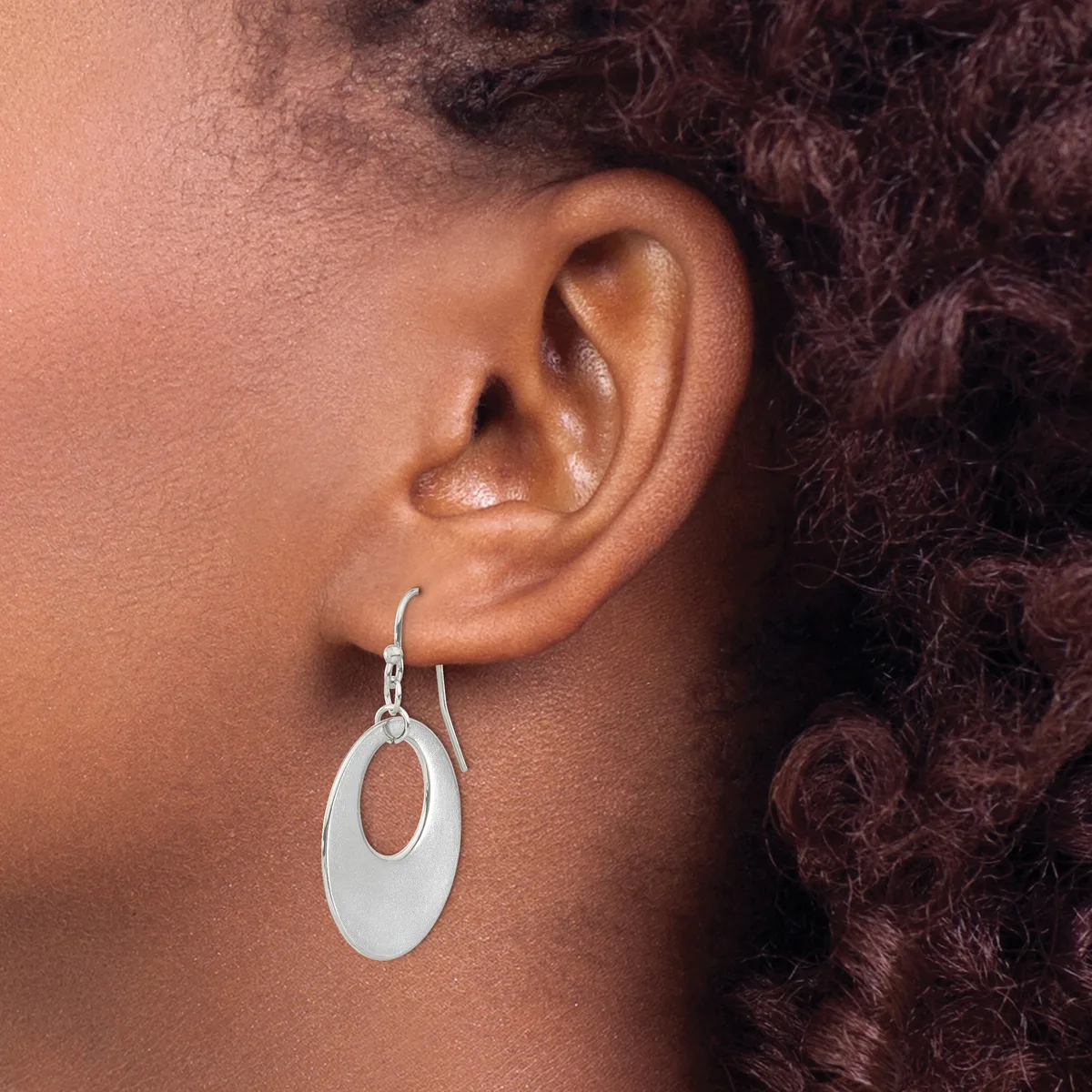 A person wearing a silver oval drop earring with an open center, shown from the side view against their ear and dark curly hair.