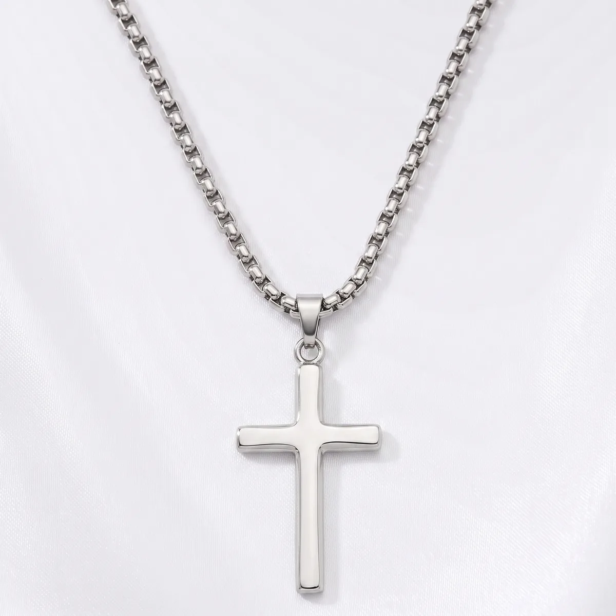 A silver cross pendant hangs from a silver box chain necklace against a white background.